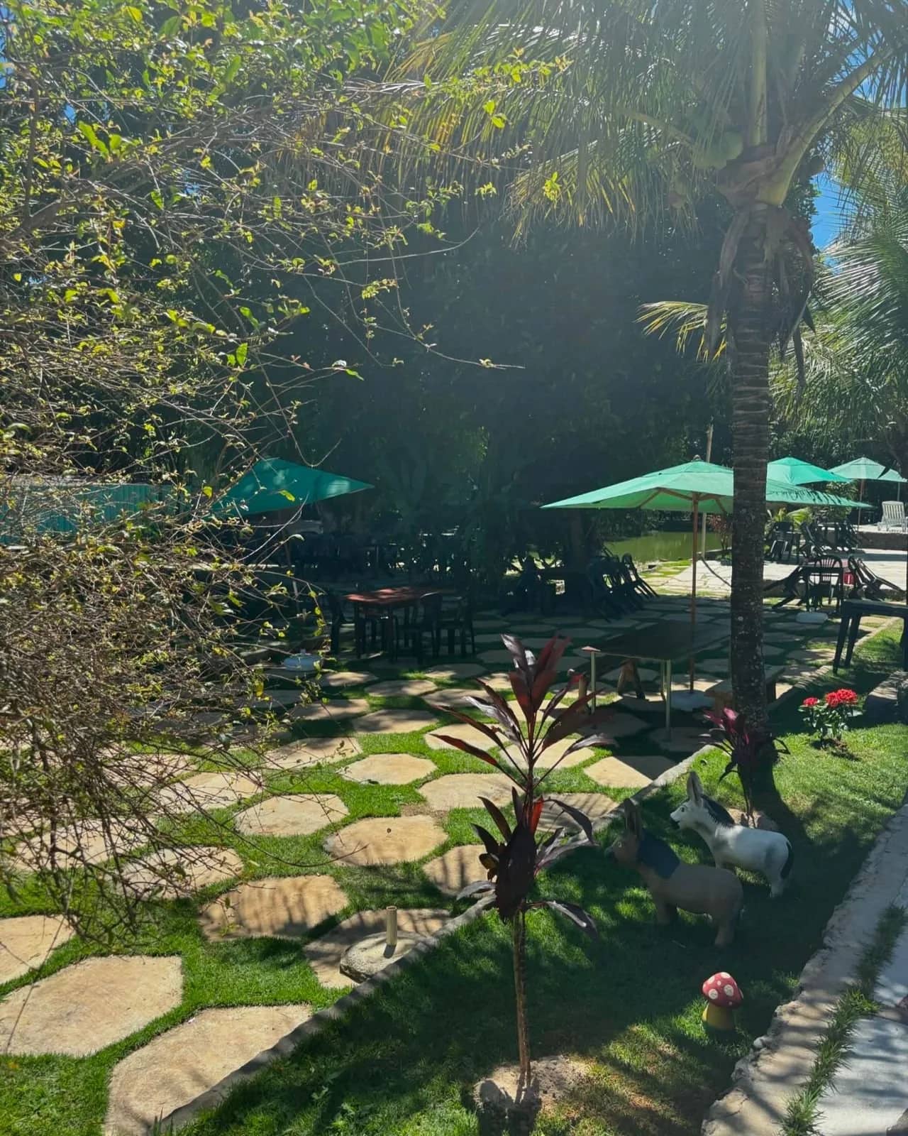 Mesas sob guarda-sóis verdes em jardim arborizado com estatuetas decorativas