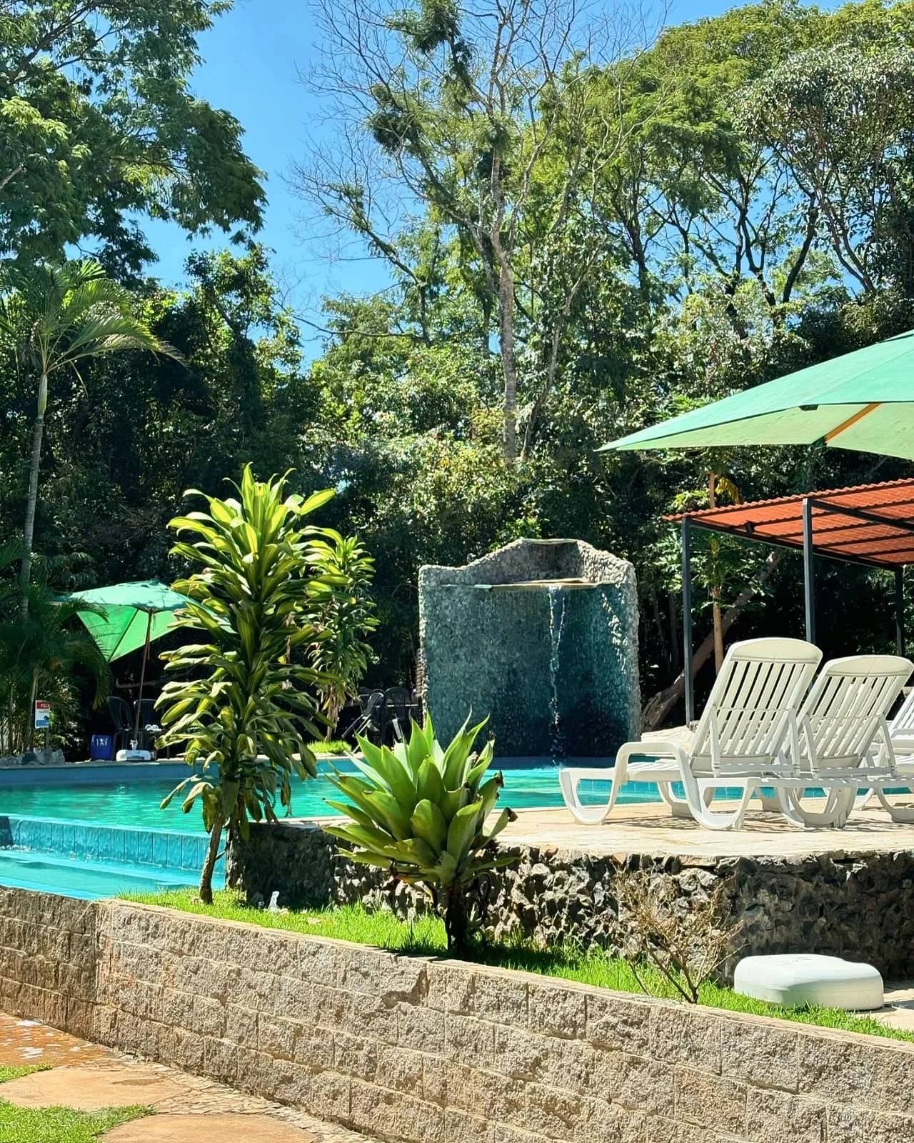 Piscina com cascata de pedra, espreguiçadeiras brancas e guarda-sol verde