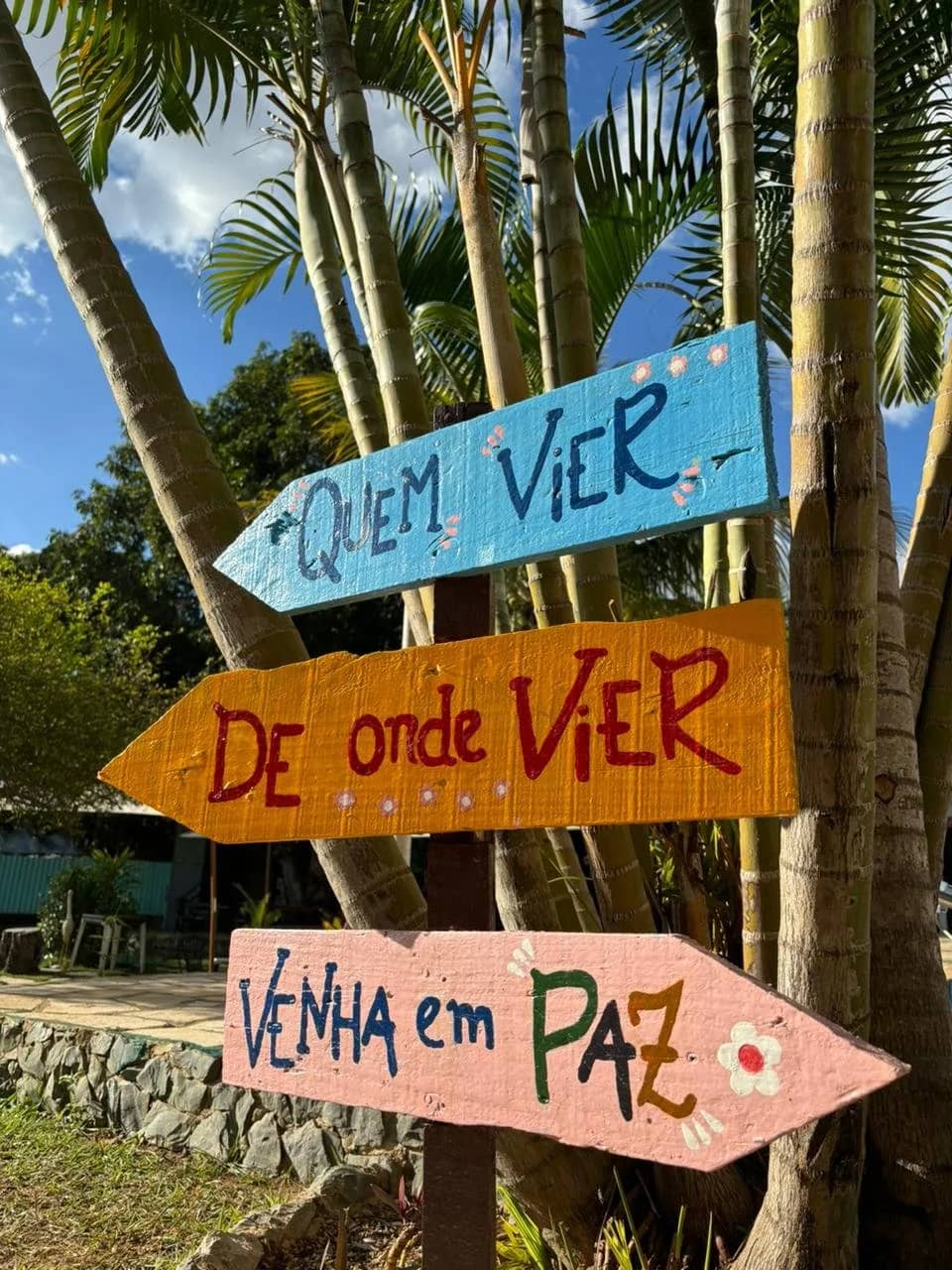 Placas de madeira pintadas à mão entre coqueiros: “Quem vier, de onde vier, venha em paz”