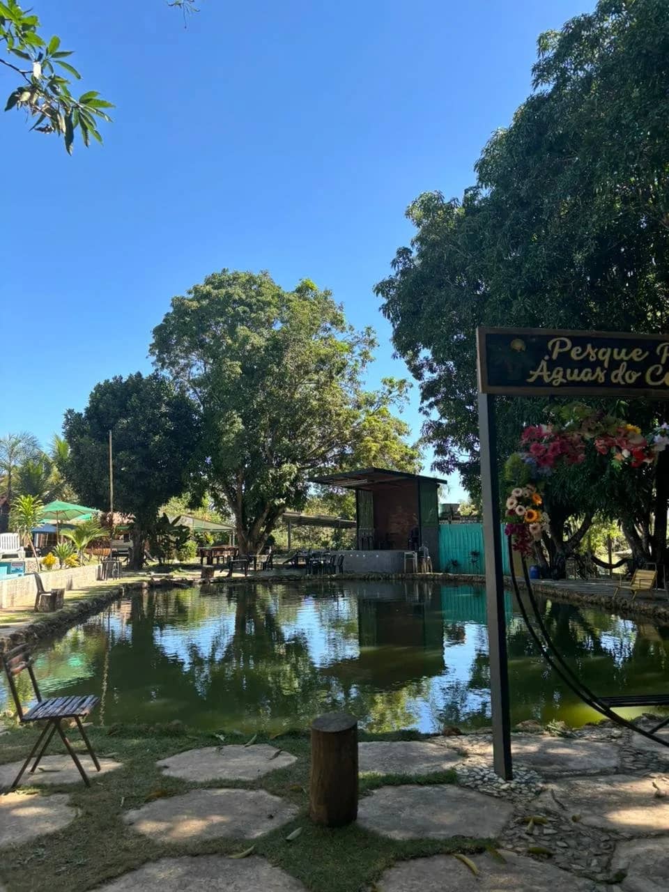 Lagoa tranquila refletindo árvores, com bancos e área de mesas na margem