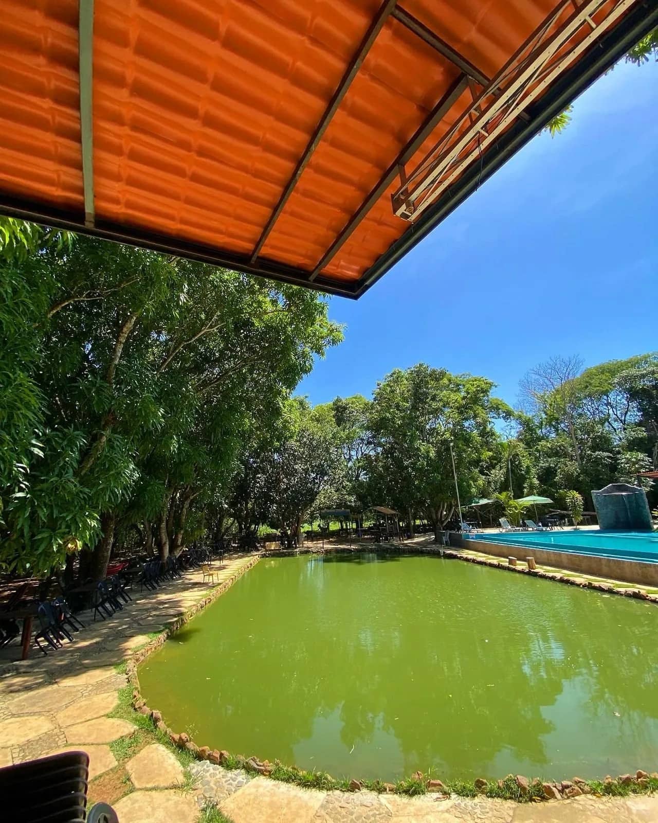 Lagoa verde vista de sob a cobertura laranja, com mesas e piscina ao lado