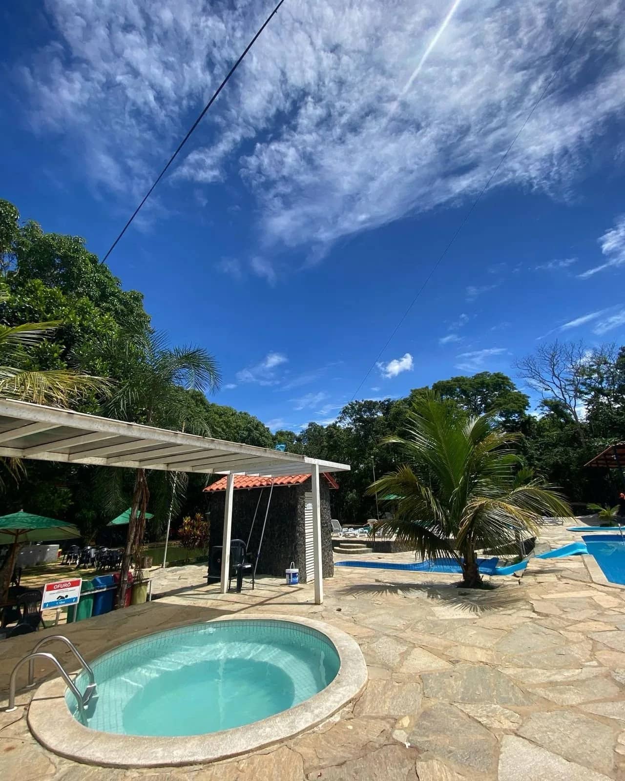 Ofurô redondo no piso de pedra sob céu de nuvens finas, piscinas ao fundo